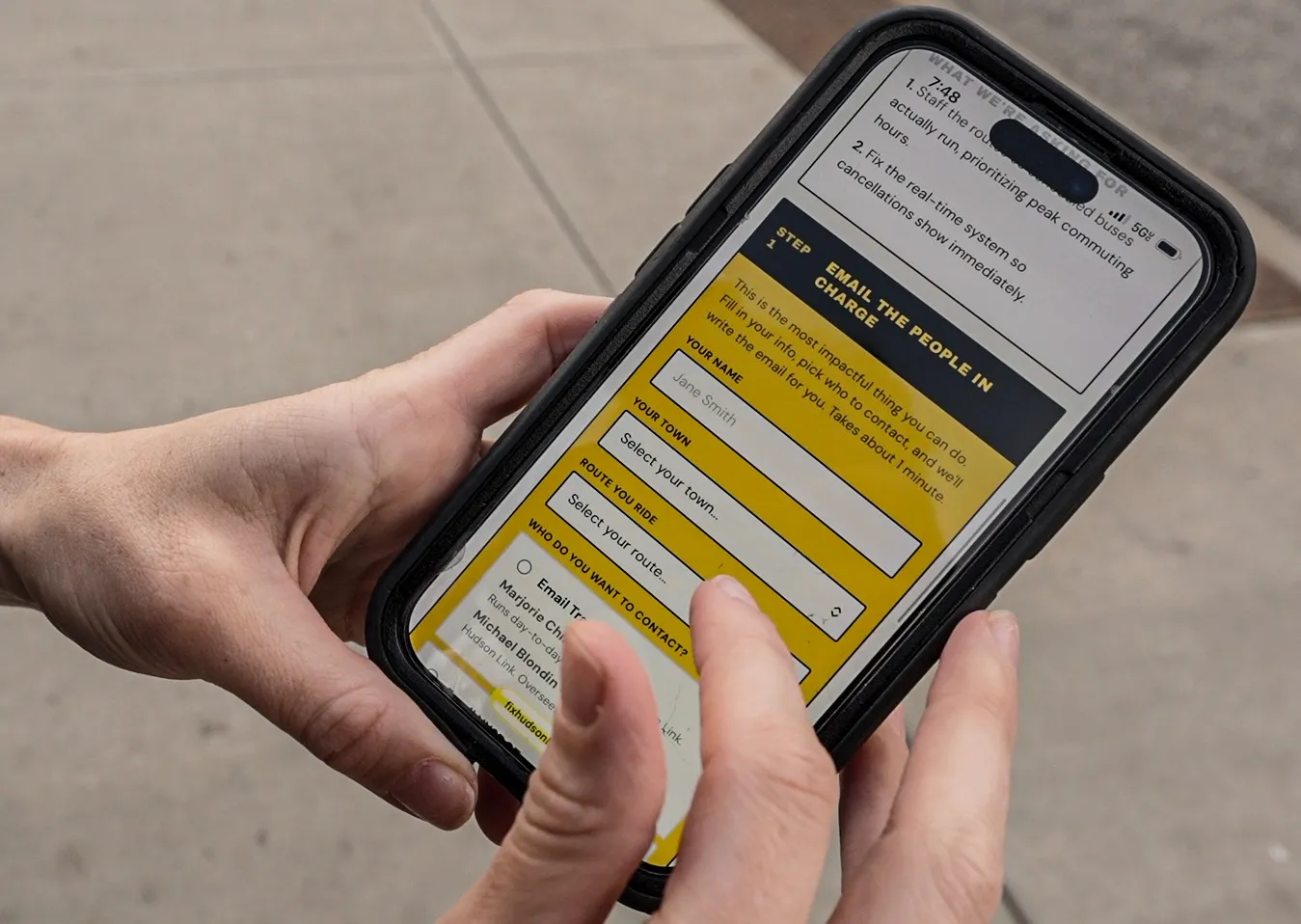 A rider holds a phone showing the fixhudsonlink.com site on a Rockland sidewalk